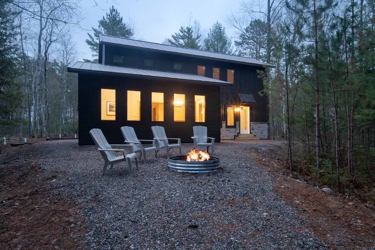 An outdoor fire pit is surrounded by several comfortable chairs, set on a gravel area. The two-story rental features large windows that reflect the natural surroundings, while tall trees frame the space, creating a serene and inviting atmosphere.