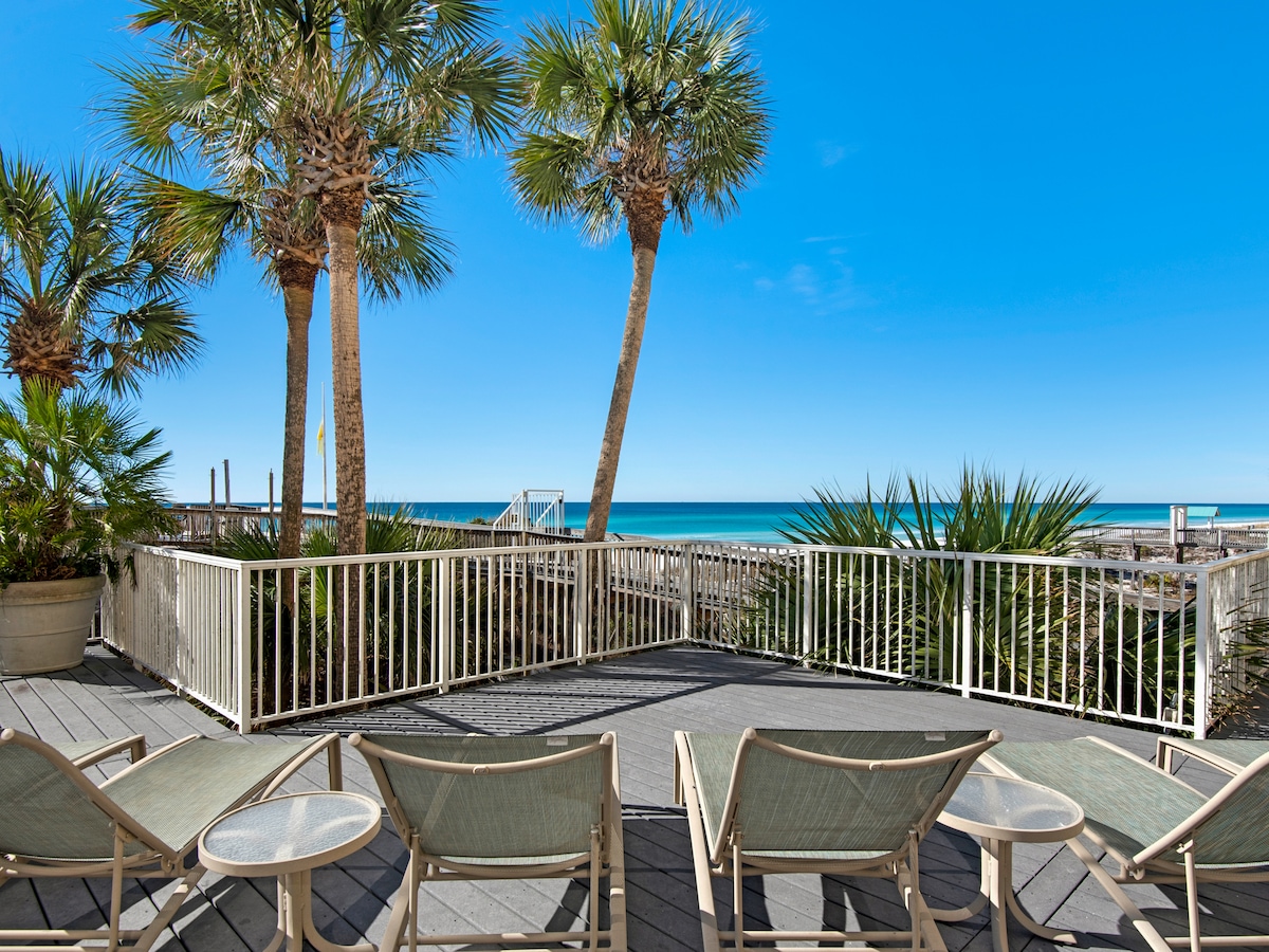 A spacious deck is displayed with several lounge chairs facing the Gulf. Lush palm trees and other greenery frame the area, complementing the unobstructed views of the turquoise water under a clear blue sky.