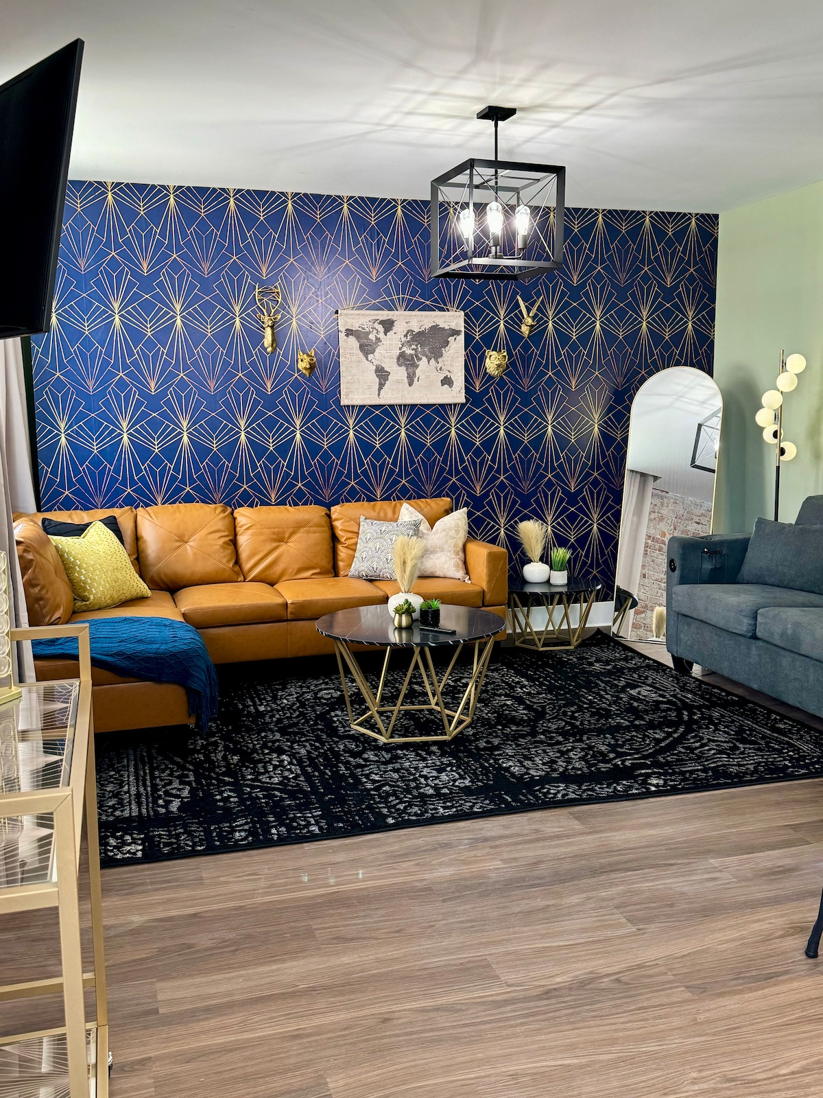 A modern living room is featured with a sleek gold-accented coffee table and a plush, caramel-colored sectional sofa. A patterned rug adds texture to the warm wooden floor. A wall with geometric designs is highlighted, complementing the softly lit area, which includes decorative accents and a large mirror.