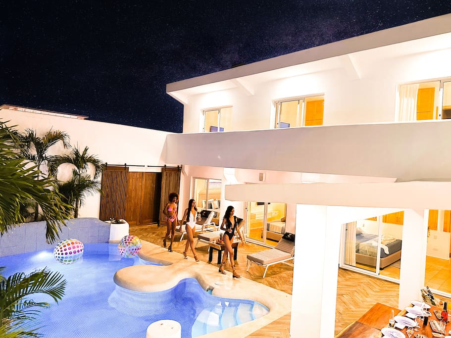 The modern villa features a luminous pool area surrounded by vibrant greenery, illuminated by soft lighting under a starry sky. Comfortable loungers are positioned beside the uniquely shaped pool, while a small gathering of people enjoys a relaxing evening.
