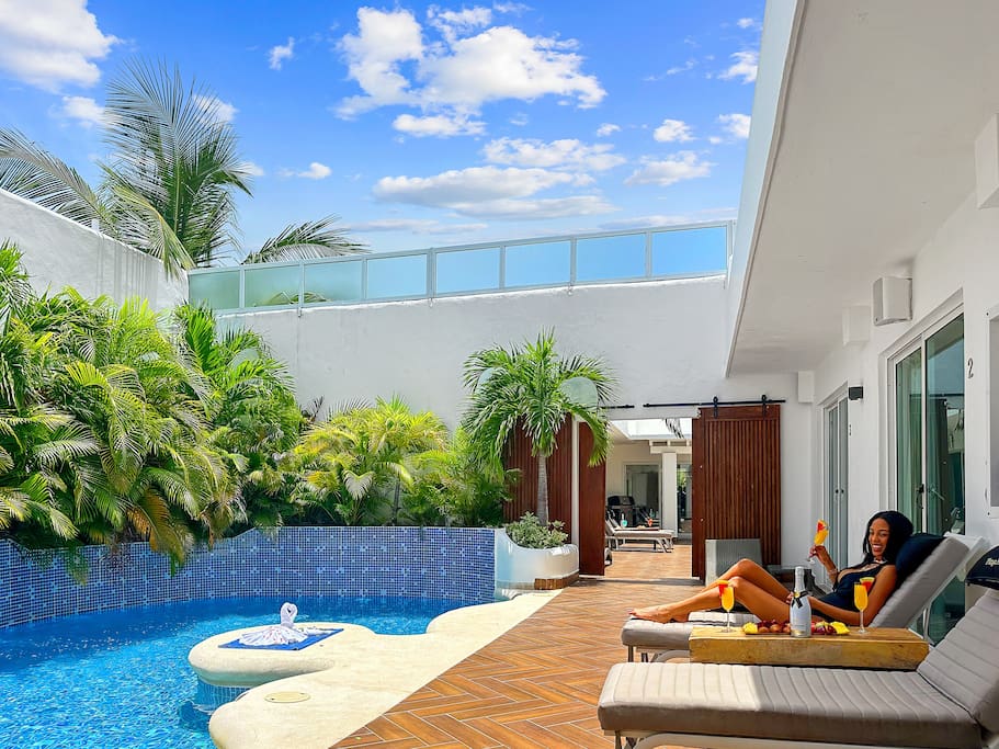 A well-appointed outdoor area features a private pool surrounded by lush greenery. Lounge chairs are positioned near the pool, inviting relaxation under a blue sky. Sliding glass doors lead to an interior space, enhancing the connection between indoor and outdoor living.