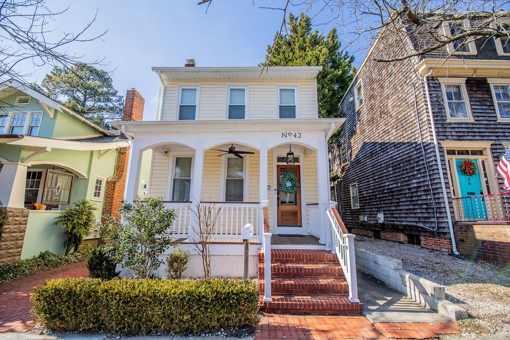 A classic two-story single-family home is presented with a light yellow exterior and white trim. The front porch features a welcoming entrance with a decorative wreath, surrounded by neatly landscaped bushes and a brick pathway. Nearby homes exhibit varied architectural styles.