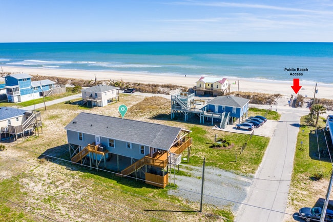 Aerial view of the property showcasing its proximity to the ocean. The beach is visible in the background, with clear blue waters and a sandy shore. A sign indicates the nearby public beach access. Nearby structures and parking areas are also visible within the scene.