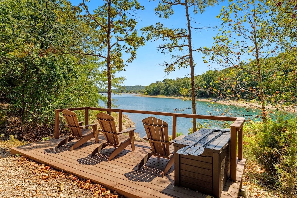 A serene lakefront deck features four Adirondack chairs positioned for enjoying views of the water. A wooden storage box sits nearby, surrounded by lush trees that provide a natural backdrop to the tranquil setting.