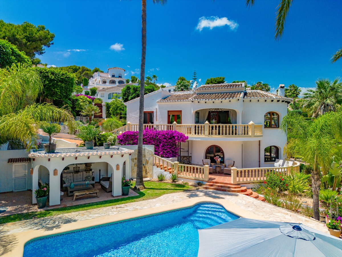 The villa features a two-story design surrounded by lush greenery and vibrant bougainvillea. An inviting swimming pool is set in the foreground, with a shaded lounging area nearby. A sunny terrace provides ample space for relaxation, enhancing the charm of the property.