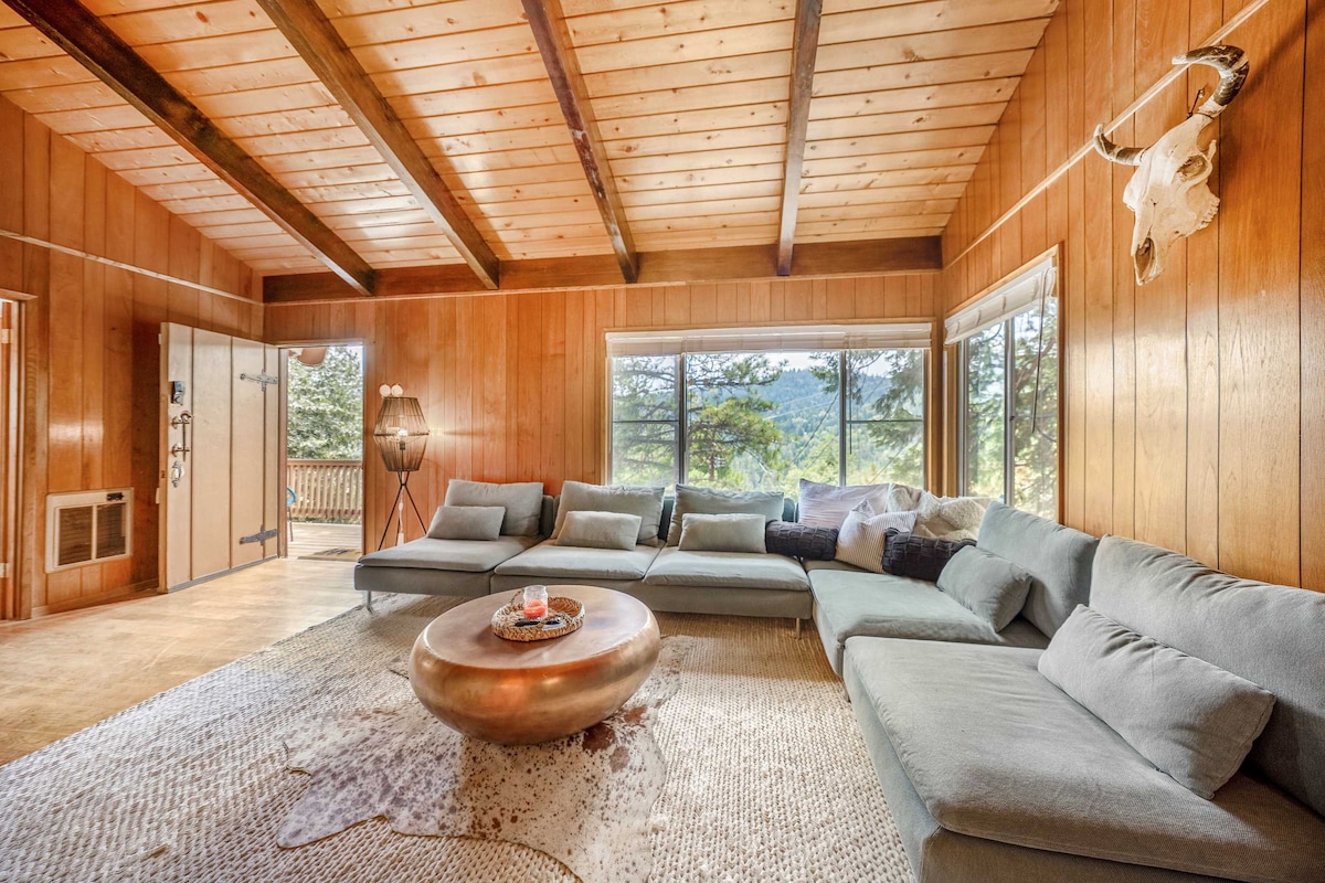 A spacious living area features a large sectional sofa arranged around a round coffee table. Natural light enters through expansive windows that provide mountain views. Warm wood paneling and a beamed ceiling create a cozy and inviting atmosphere.
