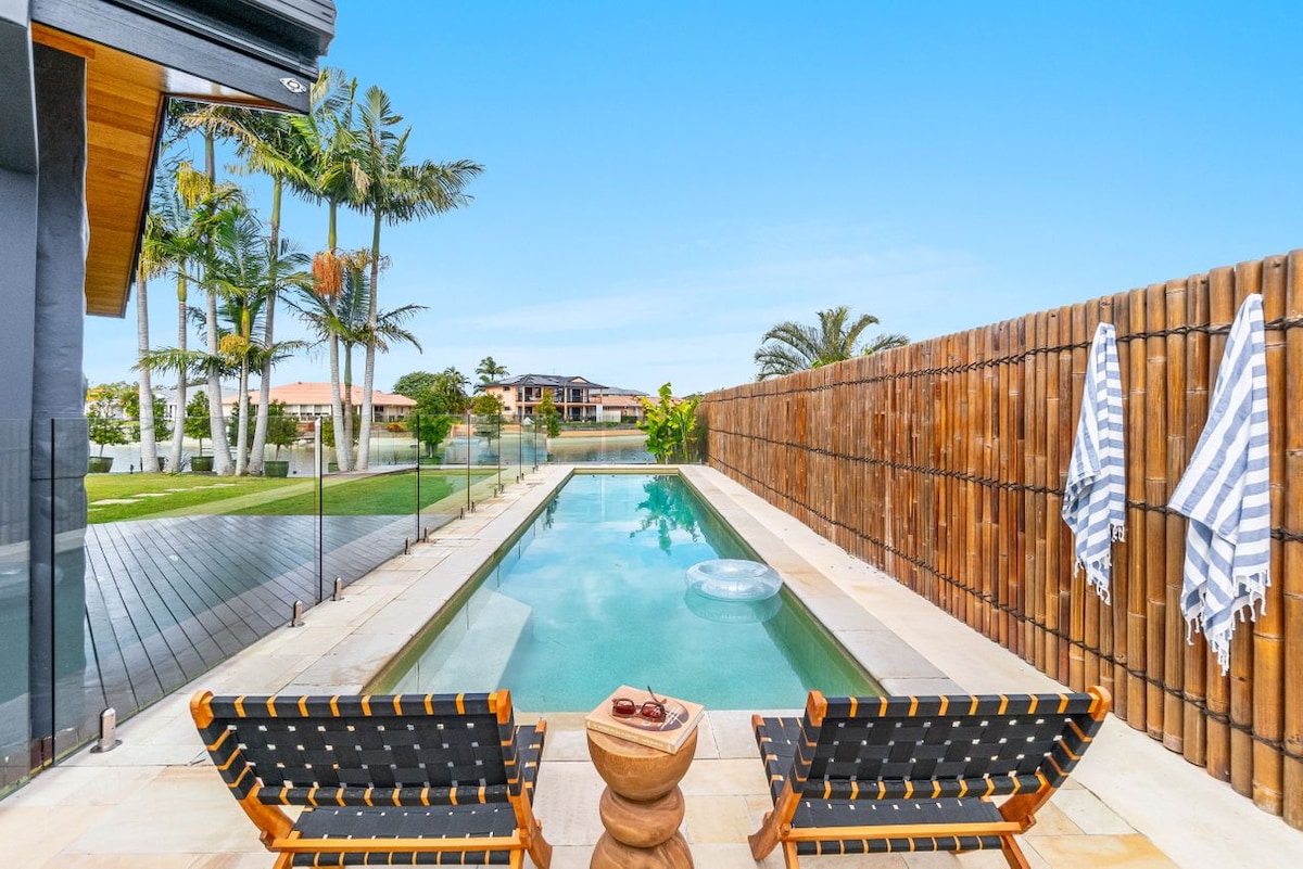 A serene outdoor area features a tranquil swimming pool surrounded by palm trees, with two lounge chairs positioned for relaxation. A wooden fence offers privacy, while towels hang neatly nearby. The clear blue sky enhances the peaceful ambiance of the waterfront setting.