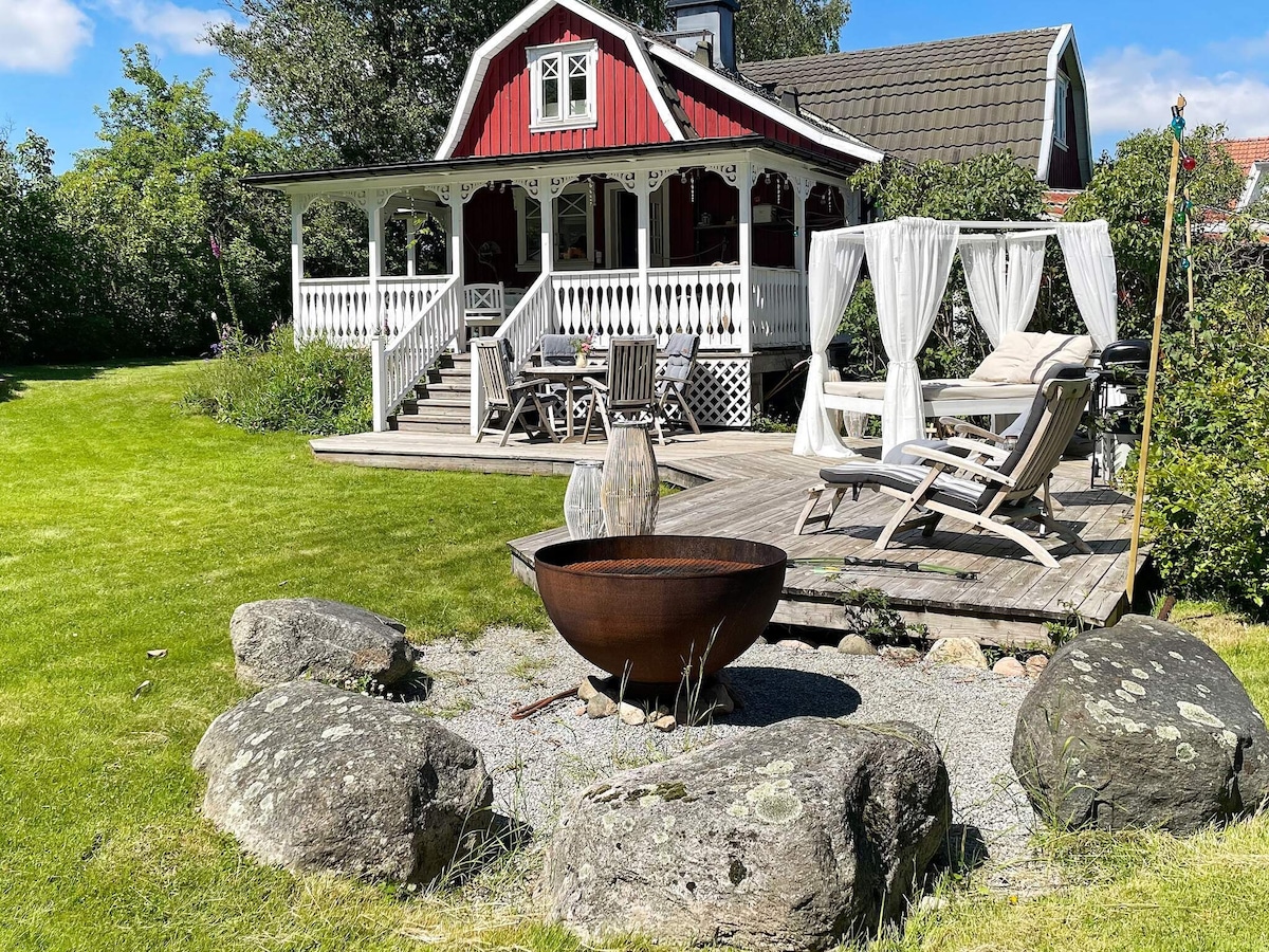 A charming exterior view of a red cottage surrounded by a lush green garden. A wooden deck with lounge chairs and a dining area is visible, alongside a cozy seating area draped in white. A decorative fire pit is set in a gravel area adorned with large stones.