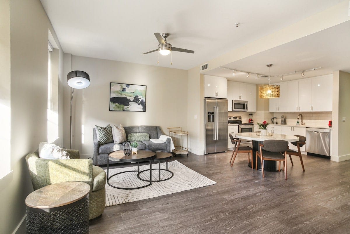 A modern living space features a comfortable seating area with a gray sofa, green chair, and a round coffee table. A dining table with chairs is positioned near the kitchen, which includes stainless steel appliances. Soft natural light streams through large windows, highlighting the open layout.