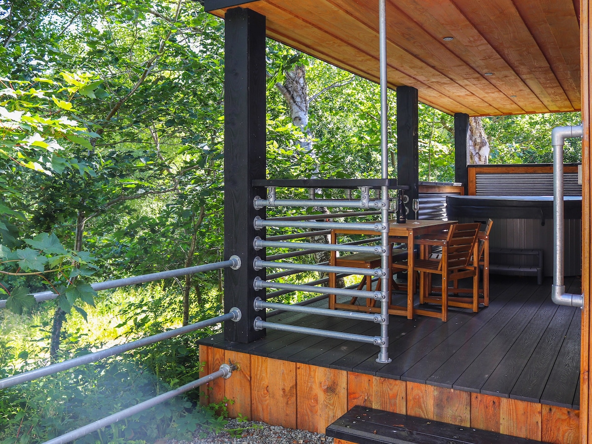 An outdoor seating area is visible, featuring a wooden dining table and chairs set against a backdrop of lush greenery. The structure features wooden flooring and vertical wooden posts, while metal railings provide a contemporary touch.