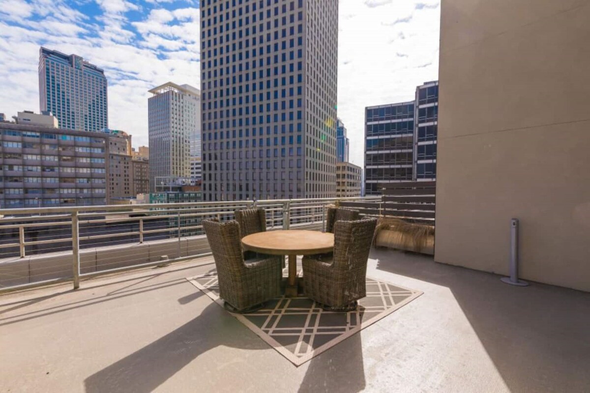 A rooftop terrace features a round dining table surrounded by four wicker chairs. The backdrop showcases the skyline of downtown New Orleans, with modern high-rise buildings under a partly cloudy sky.