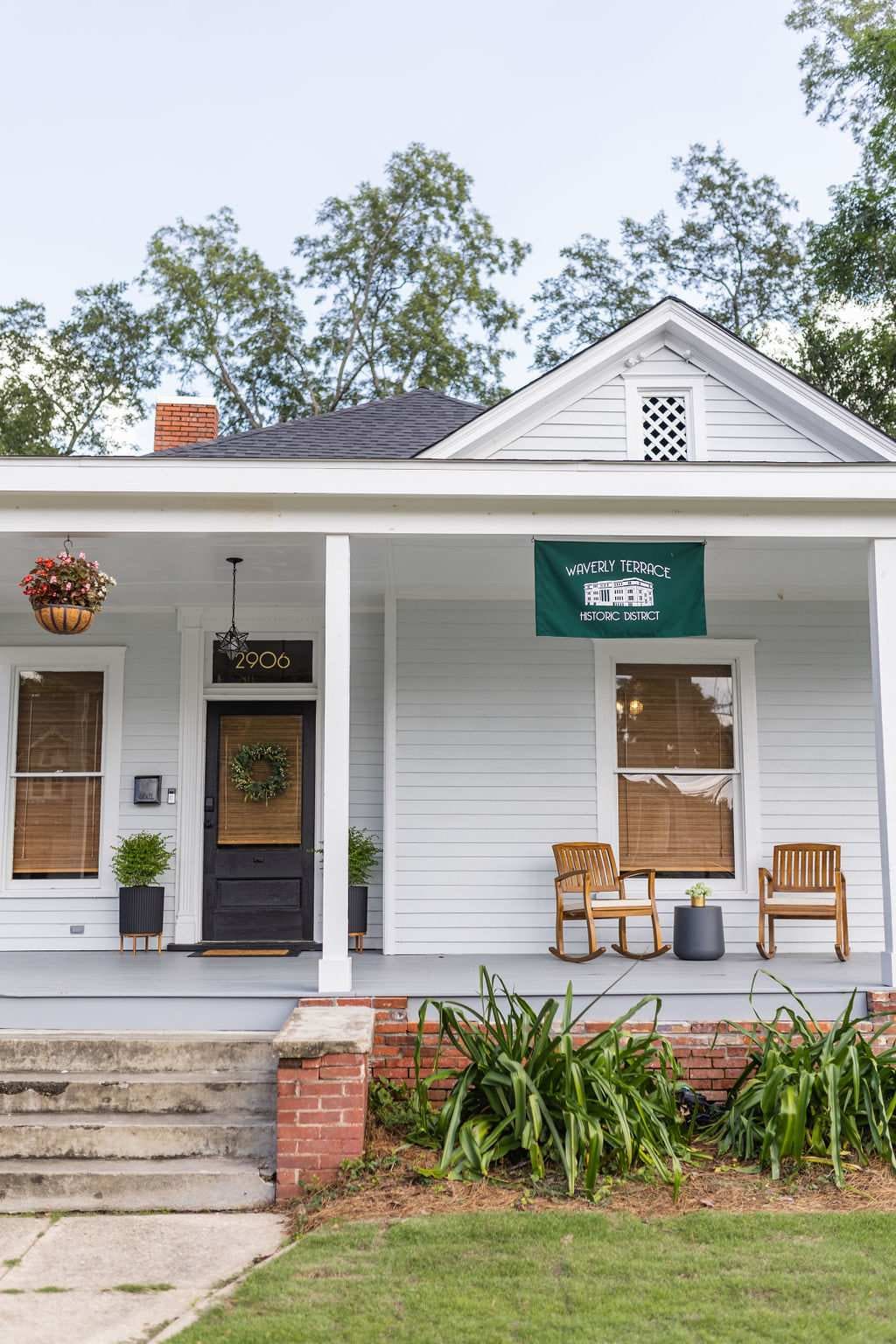 The charming exterior of the bungalow is shown, featuring a welcoming front porch with two wooden chairs and a potted plant. A green banner hangs above the door, and lush greenery surrounds the entrance.