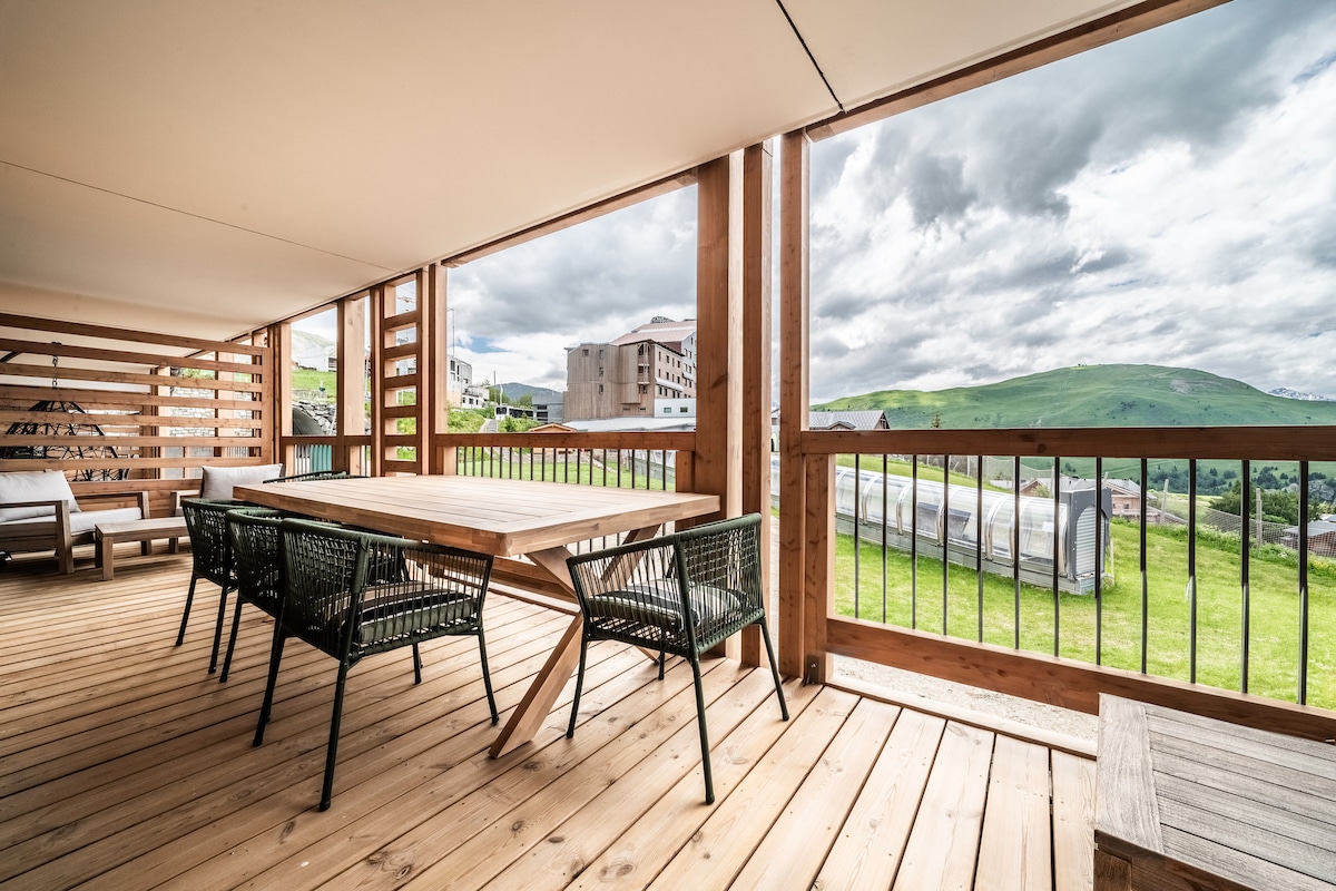 An open balcony features a wooden floor and a dining table surrounded by six black woven chairs. A spacious seating area with light cushions is present, offering views of the green hills and cloudy sky through wooden railings.