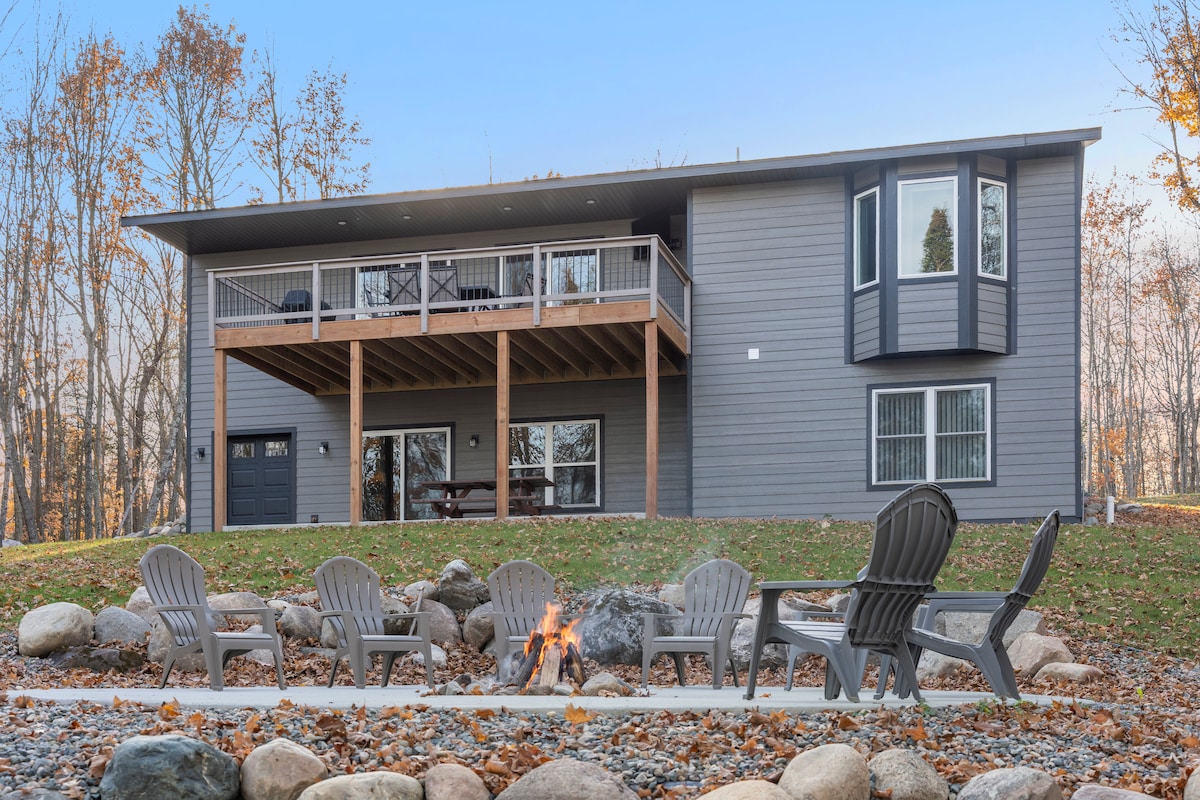 The exterior of the cabin is displayed with a spacious deck accessible from multiple exits. A fire pit surrounded by several Adirondack chairs is visible in the foreground, offering a cozy gathering spot. The surrounding trees exhibit autumn colors, adding to the natural setting.