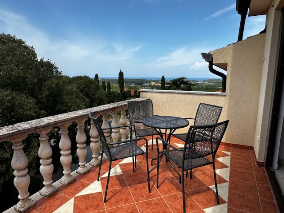 A private balcony features a round table surrounded by four chairs, set against a backdrop of green trees and distant hills. The terrace floor is tiled, with a decorative pattern enhancing the spacious feel, while the railing is adorned with stone balusters.