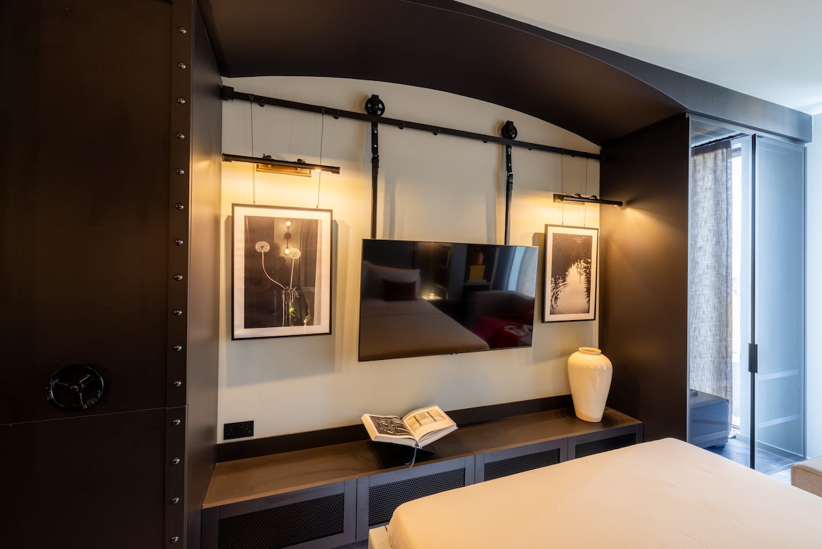 A stylish and modern studio features a wall-mounted flat-screen TV above a sleek, low console table. Two framed artworks are displayed on the wall, accompanied by contemporary wall sconces. A decorative vase sits atop the console, with natural light filtering in through the large window.
