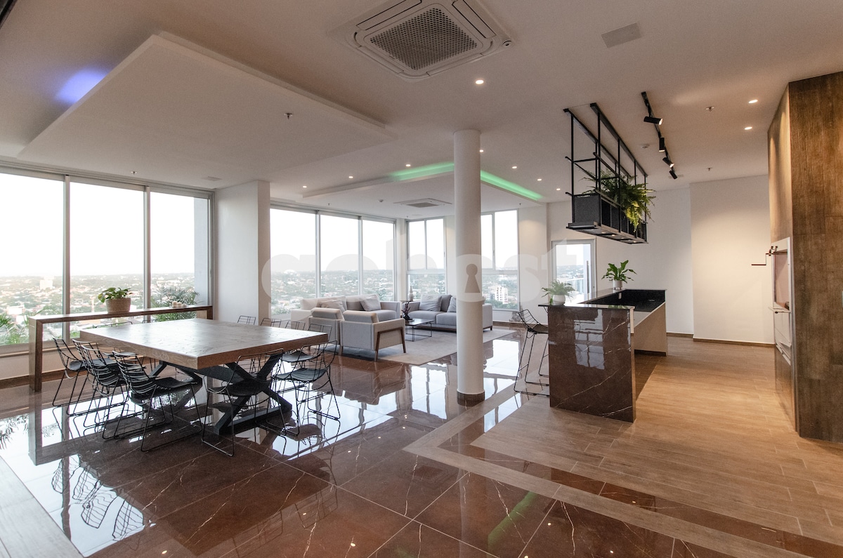 An open living space features large windows offering views of the surrounding area. A dining table for six is positioned prominently, while a kitchen area with modern fixtures is visible. Soft seating is arranged in the living room, providing a comfortable setting.