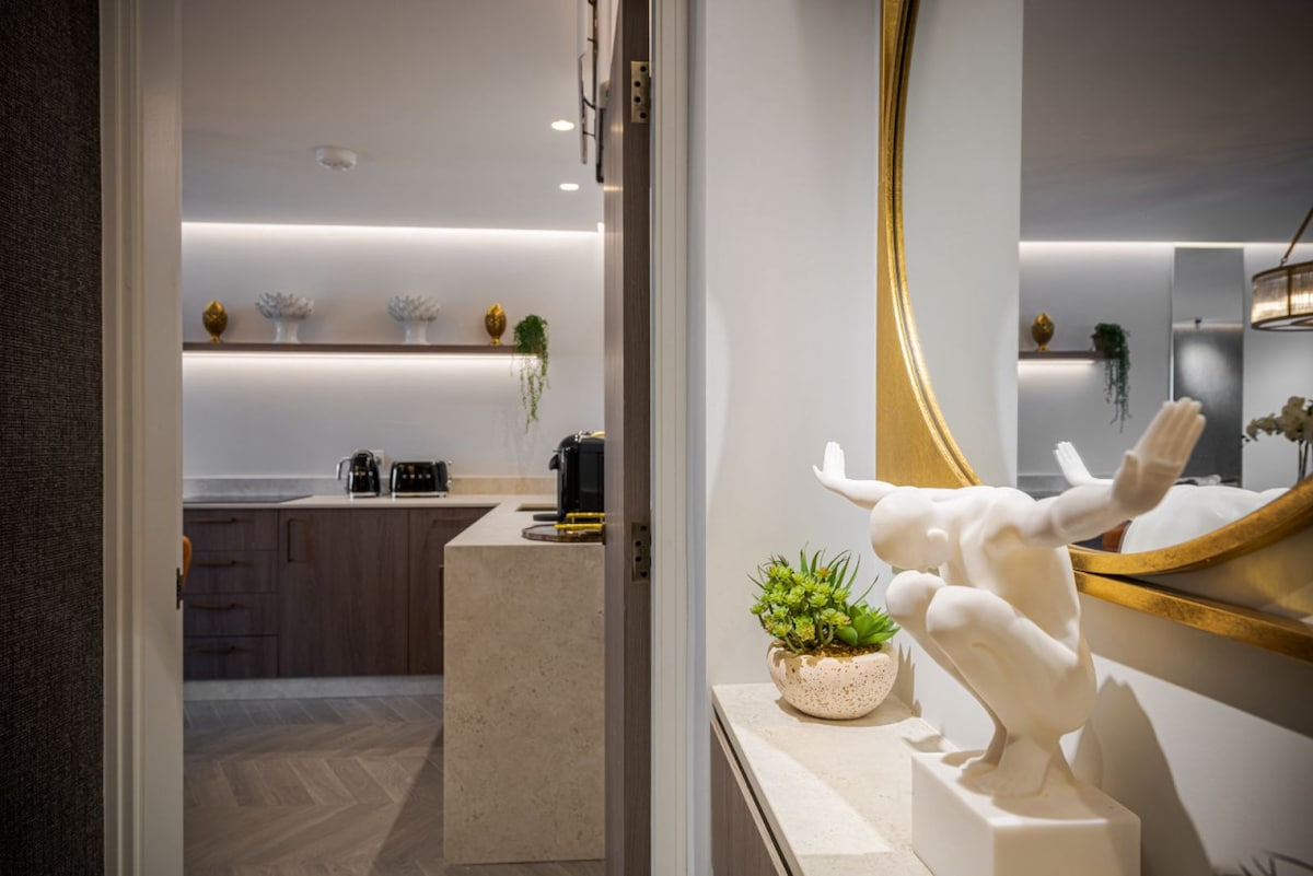 A modern entryway showcases a sculptural figure and a small potted plant atop a sleek surface. The background reveals a well-equipped kitchen with elegant cabinetry and soft lighting, creating a refined atmosphere.