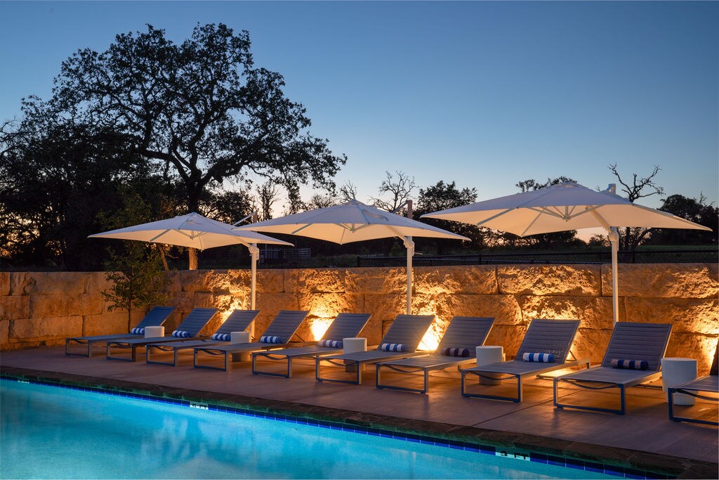 A serene pool area is highlighted by elegant lounge chairs arranged beneath large white umbrellas, offering shade. Soft lighting enhances the stone walls, creating a calming ambiance as dusk settles over the Texas landscape.