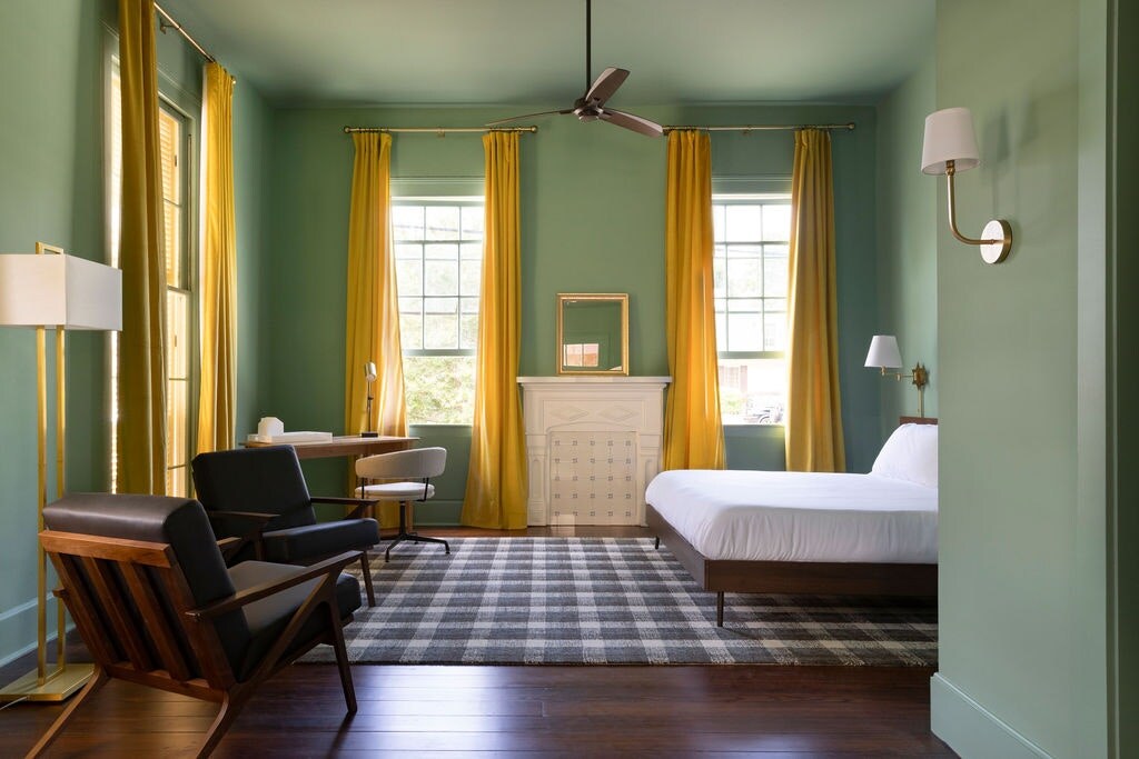 A bright room features green walls accompanied by tall yellow curtains framing large windows. A white bed with a simple design is positioned on a checkered rug. A stylish chair and a small desk complete the space, with a decorative fireplace adding character.