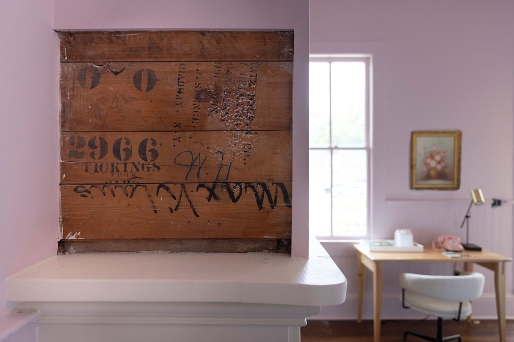 A close-up of an antiqued wooden wall with faint markings and remnants of old designs. To the right, a softly lit workspace features a simple desk with a chair, a decorative wall art piece, and a window allowing natural light to fill the space.