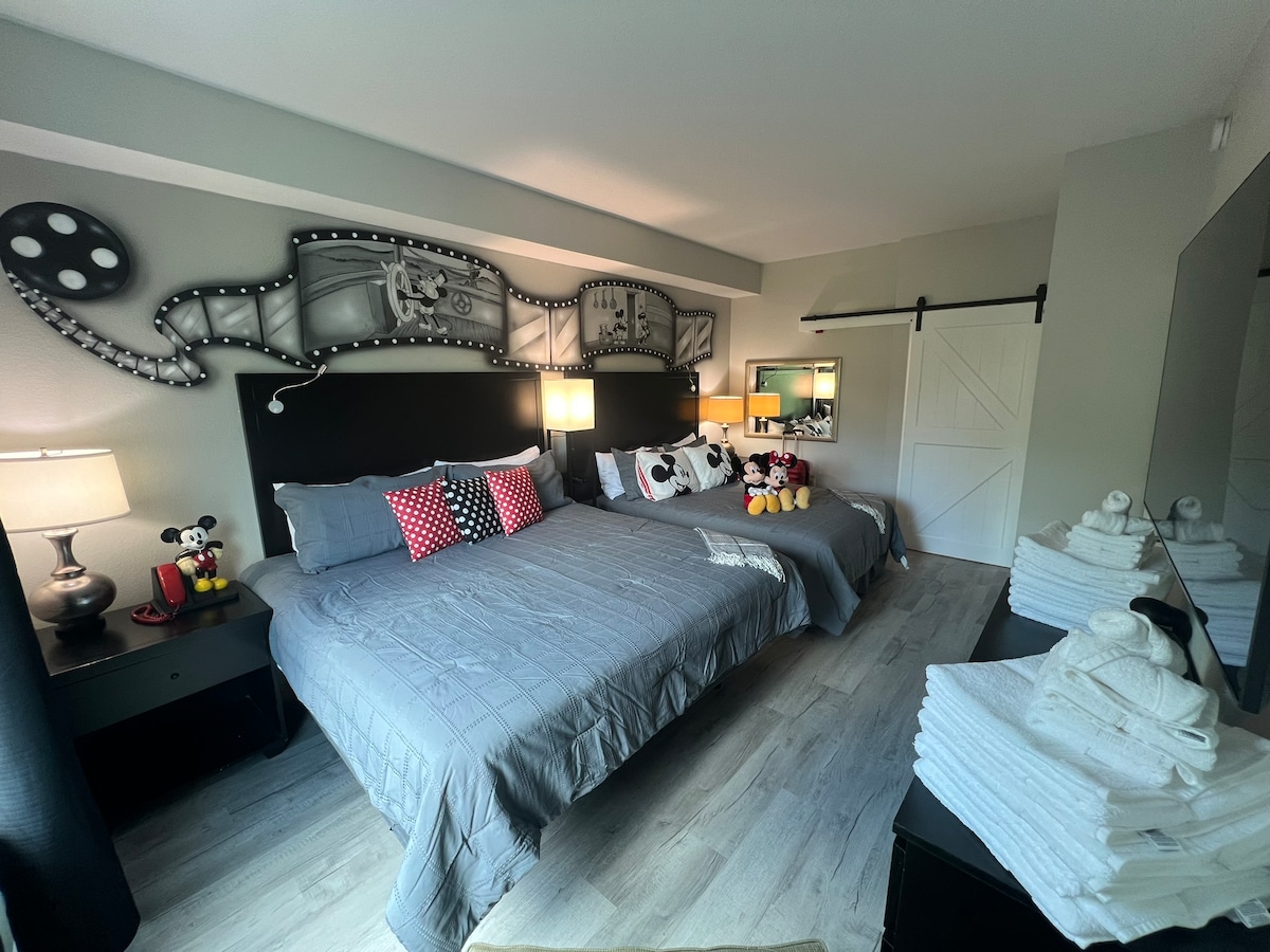 The sleeping area features two spacious beds accompanied by decorative pillows and stuffed toys. Nightstands with lamps provide soft illumination. A large mirror reflects the room's design, which is enhanced by a wall mural inspired by classic cinema.