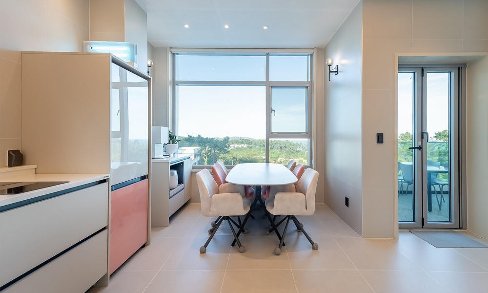 A modern kitchen area displays sleek cabinetry in soft pastel colors. A spacious dining table is surrounded by comfortably cushioned chairs, with large windows offering a view of the surrounding landscape. Natural light brightens the room, emphasizing the clean lines of the design.