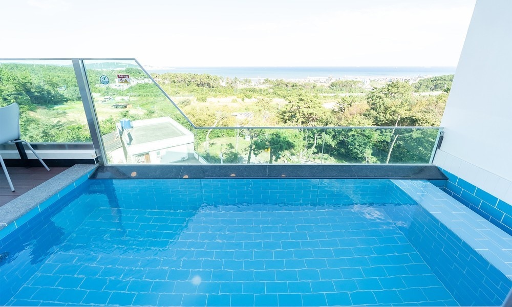 A private swimming pool is situated on a balcony with a glass railing, offering expansive views of the surrounding greenery and the ocean in the background. The pool features bright blue tiles, contributing to a serene atmosphere.
