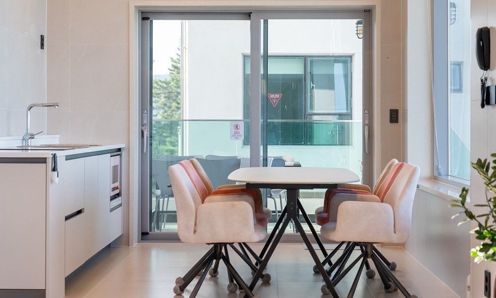A bright dining area is presented with a white table surrounded by four comfortable chairs featuring soft upholstery. Large sliding glass doors lead to an outdoor balcony, allowing natural light to fill the space while offering views of the surroundings.