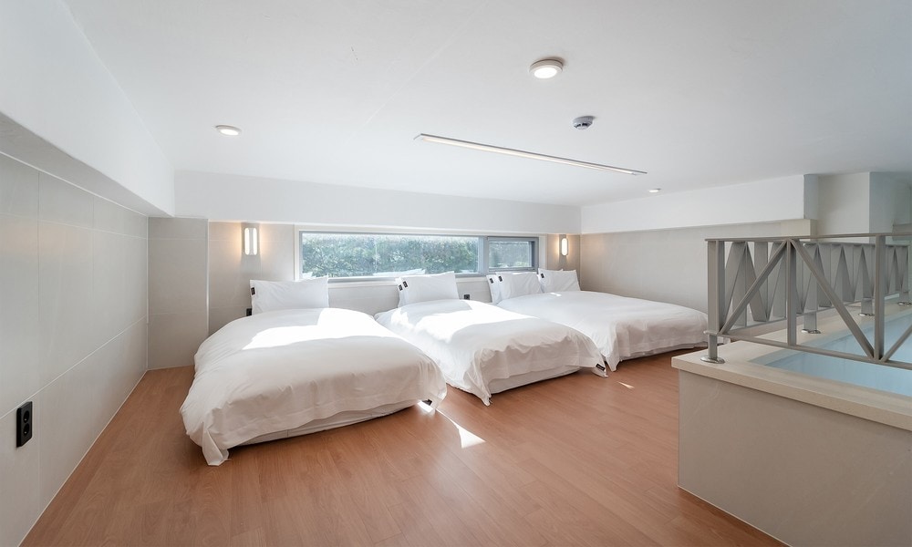 The spacious sleeping area features three beds with white linens, creating a clean and serene environment. Natural light streams through a large window, illuminating the room, while light-colored wooden flooring complements the modern design.
