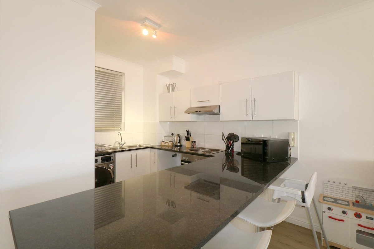A modern kitchen with sleek white cabinetry and dark countertops is shown. A stainless steel stove, microwave, and sink are visible, along with a washing machine. High stools are positioned at the countertop, creating a casual dining area. Light streams in from a nearby window.