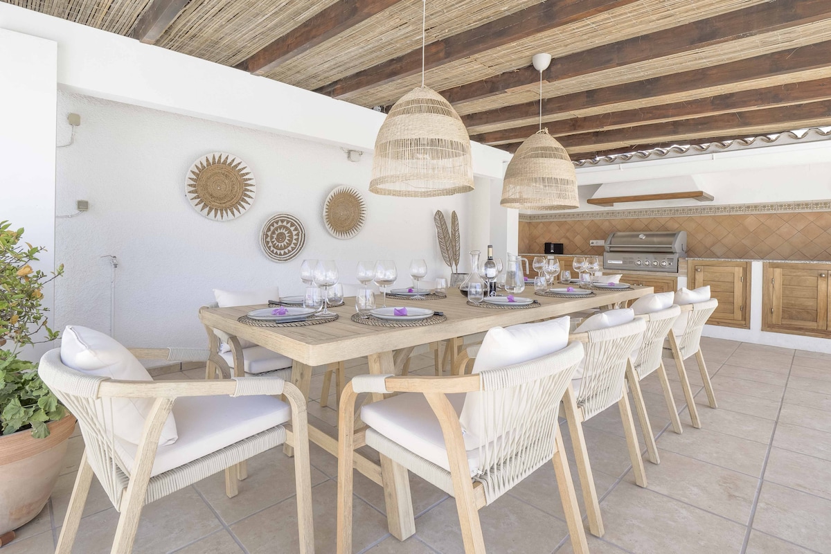 An outdoor dining area features a large wooden table surrounded by ten beige chairs, each with white cushions. Two large, woven pendant lights hang above the table, and a built-in barbecue and kitchen area are visible in the background. Decorative wall pieces add a charming touch.