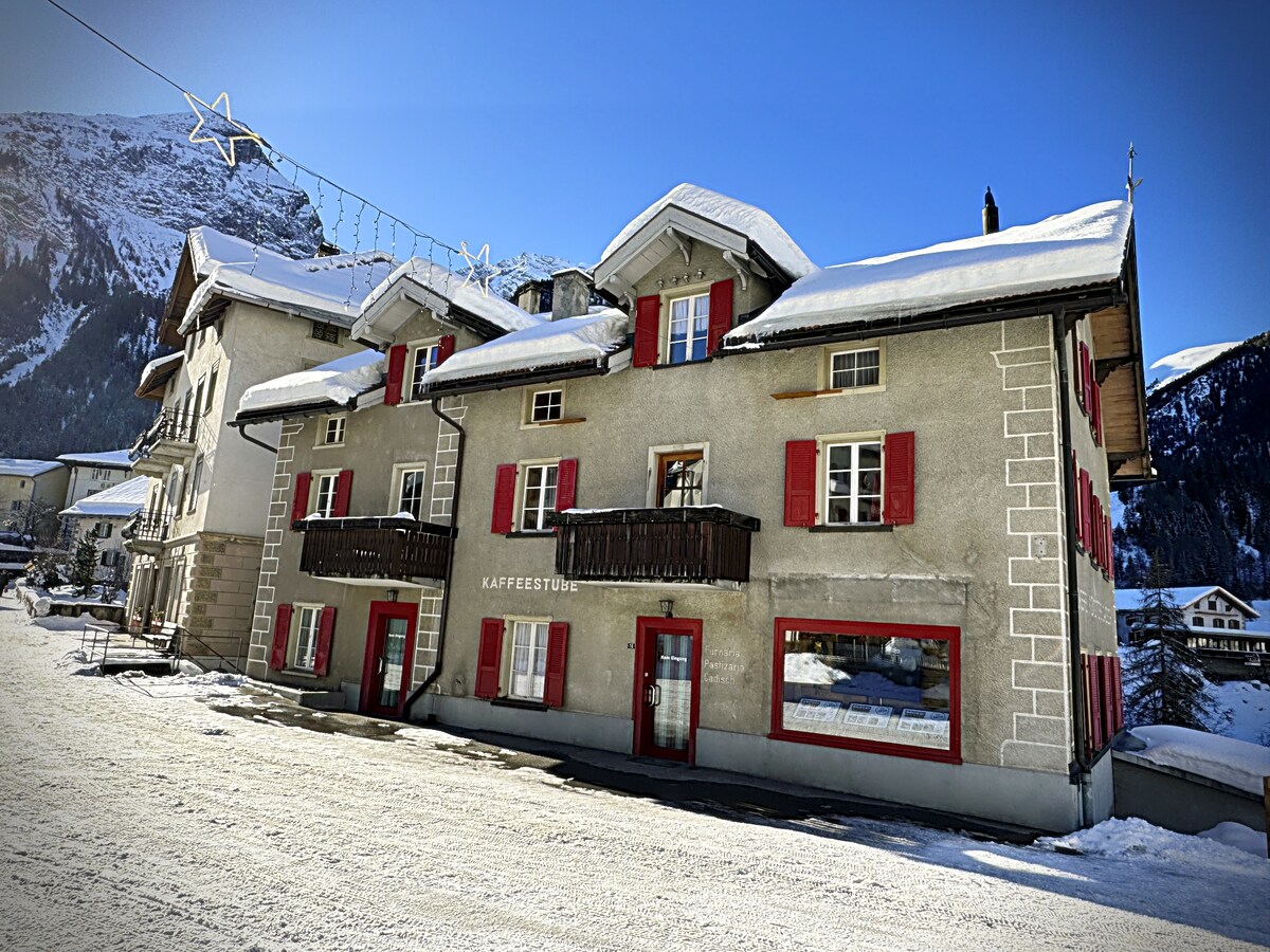 A charming building stands in a snow-covered landscape, featuring multiple levels and distinctive red shutters. The façade showcases a traditional design with a mix of balcony and large windows, inviting natural light. Snow blankets the path leading to the entrance, creating a serene atmosphere.