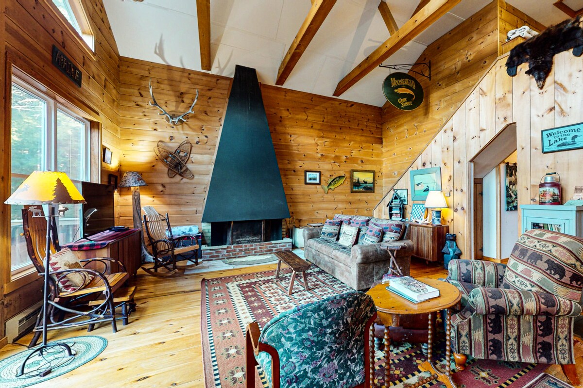 A cozy living room showcases a stone fireplace framed by wood paneling. Natural light streams in through large windows, highlighting rustic furnishings including a sofa, armchairs, and a wooden coffee table. Exposed wooden beams adorn the ceiling, enhancing the warm ambiance.