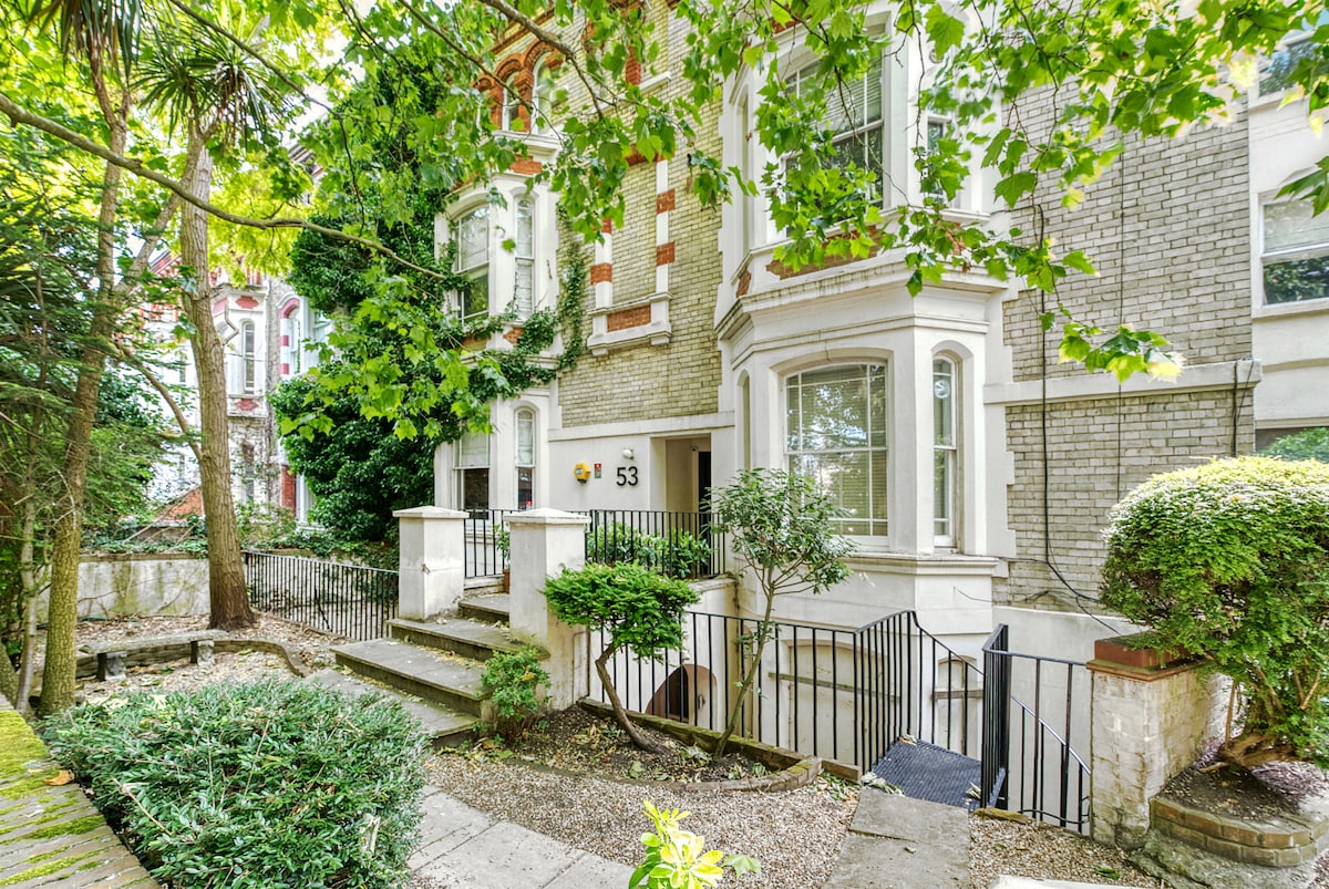 The exterior of a charming townhouse is shown, with lush greenery framing the entrance. Steps lead to the front door, and the building features large windows that permit natural light, enhancing its inviting appeal. The surrounding landscape includes manicured shrubs and trees.