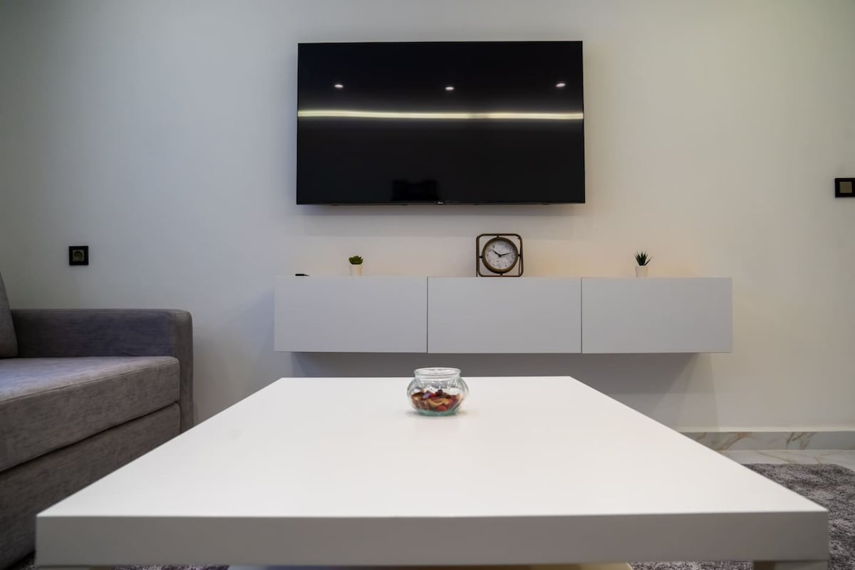 A modern living room features a minimalist coffee table at the center, with a small glass jar filled with decorative items. A sleek white cabinet is mounted on the wall beneath a flat-screen TV, providing a clean and airy feel to the space.