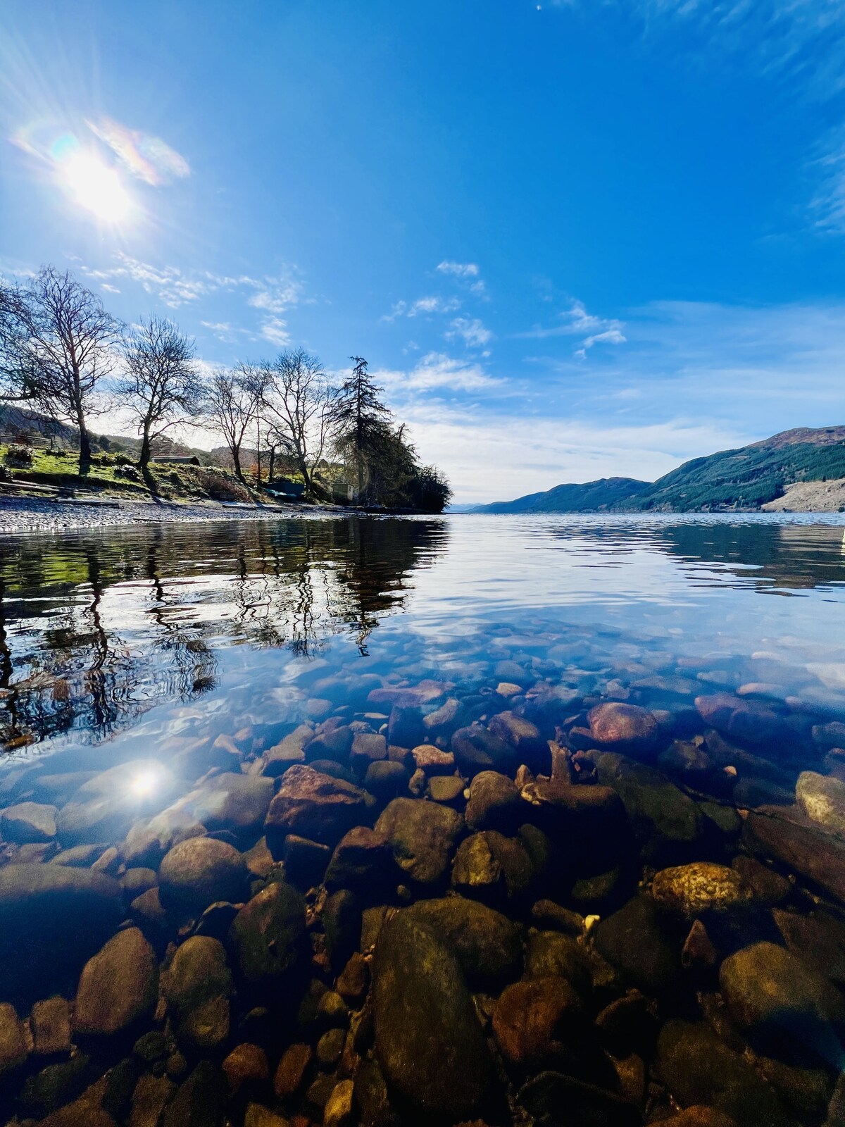 The calm surface of Loch Ness reflects the clear blue sky, with sunlit patches shimmering across the water. Smooth stones and pebbles can be seen beneath the transparent water, surrounded by lush greenery and trees along the shoreline.