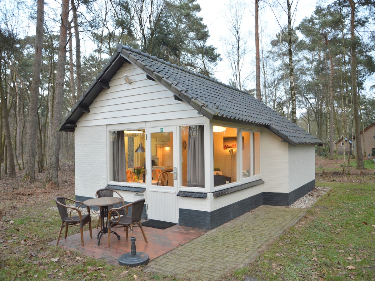 A detached bungalow is surrounded by a serene natural setting. The exterior features a sloped roof and large windows, showcasing a warm glow from within. A small terrace with two chairs is placed outside, inviting relaxation amid the quiet landscape.