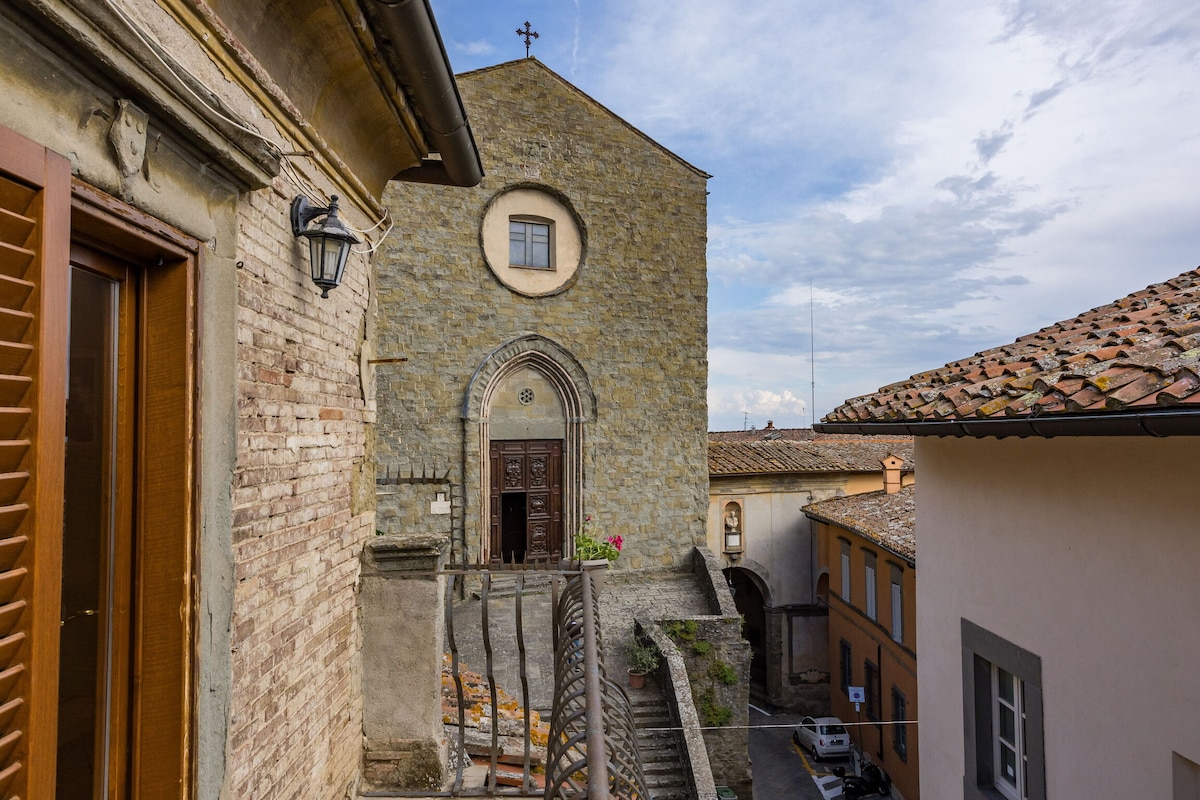 A view of the stone church facade is visible, featuring arched doorways and a circular window. Outdoor stairs lead to the church's entrance, surrounded by narrow pathways and rustic buildings. A sky filled with soft clouds adds a serene backdrop to the historical scene.