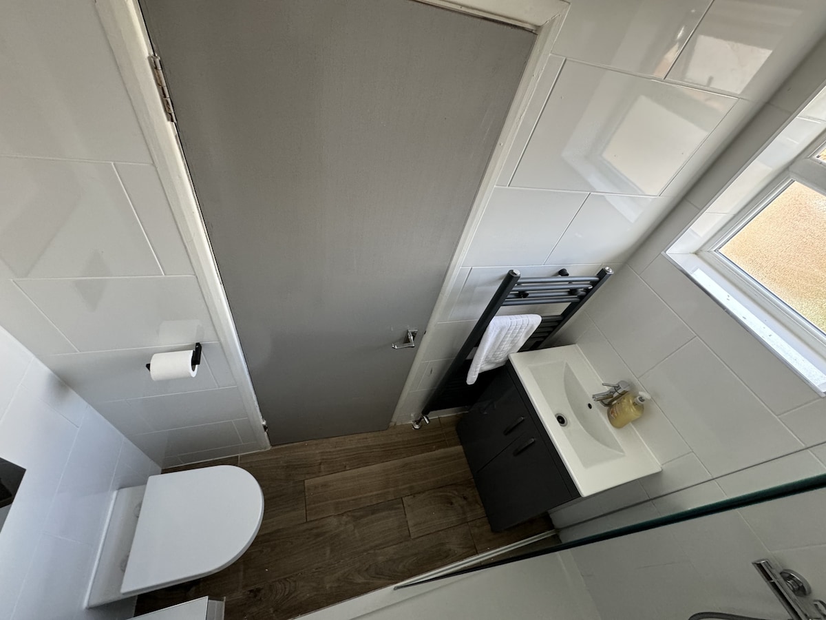 A compact ensuite bathroom is presented, featuring a modern toilet and a sleek sink. Light-colored tiles create a clean look, while a dark cabinet adds contrast. A towel rail is visible, and a window allows natural light to fill the space.