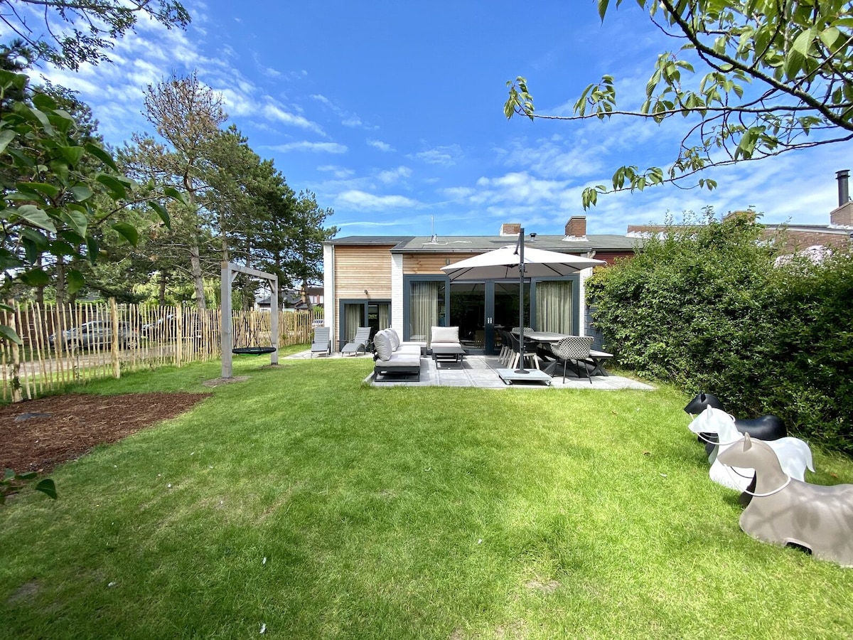 A spacious garden area features well-maintained grass, surrounded by leafy trees. Comfortable lounge chairs and a dining set with an umbrella are set up on a stone patio, inviting relaxation and outdoor meals. A children's play area is visible in the background.