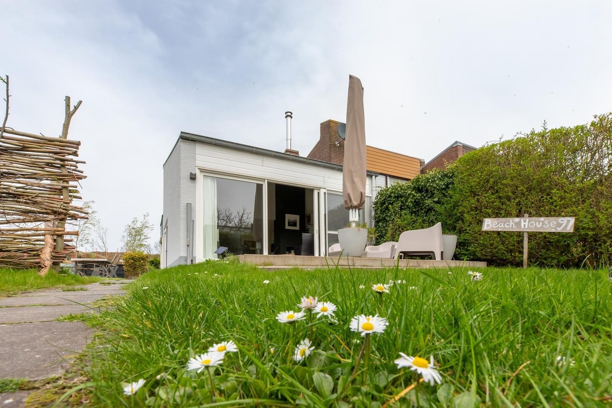 The holiday home features large glass sliding doors that offer a view of the spacious garden and terrace. A path of stone pavers leads to the entrance, with green grass and white daisies in the foreground, enhancing the charming outdoor space.