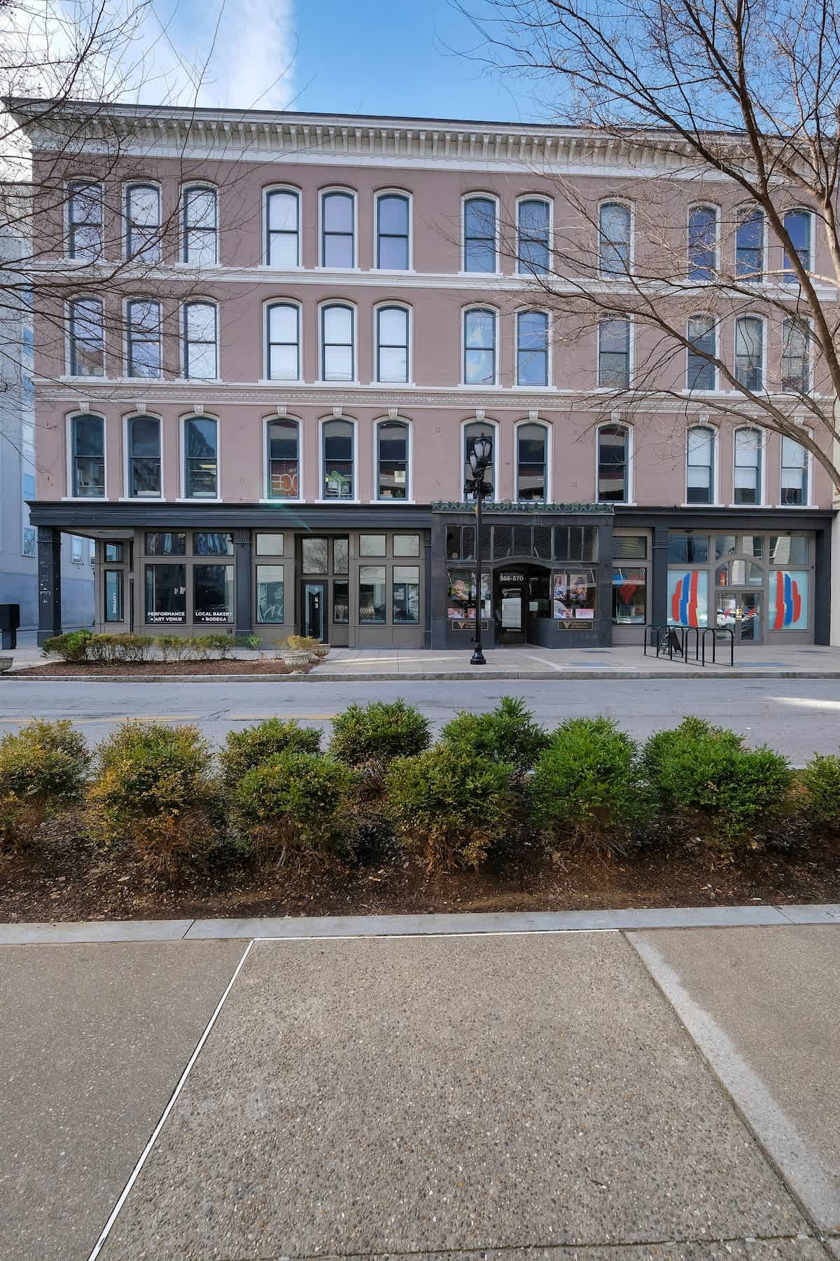 The exterior of the historic Guthrie Coke Lofts is displayed, featuring a four-story pink façade with multiple large windows. The ground level includes storefronts with glass entrances, and outdoor seating is visible along the sidewalk, surrounded by neatly maintained shrubs.