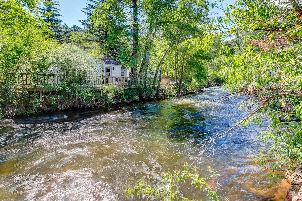 A clear view of the Big Thompson River flows gently beside the cabin, bordered by lush greenery. Sunlight reflects off the water, creating a serene atmosphere. The cabin is located on a raised platform, with a wooden deck extending towards the riverbank.