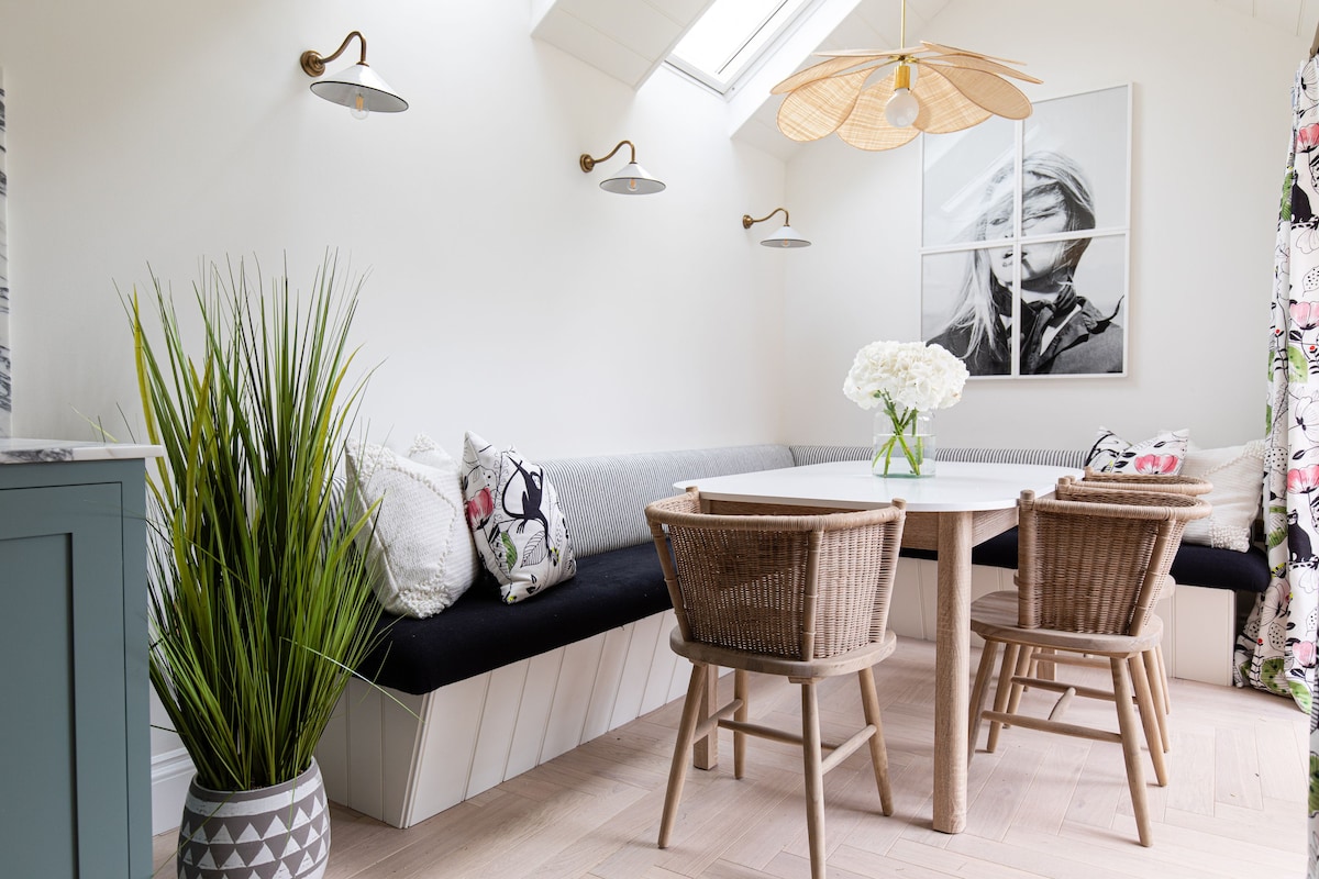 A dining area features a circular wooden table surrounded by four chairs, including a cozy banquette with plush cushions. Natural light floods the space through a skylight, while decorative wall lamps provide additional illumination. Potted greenery enhances the inviting atmosphere.