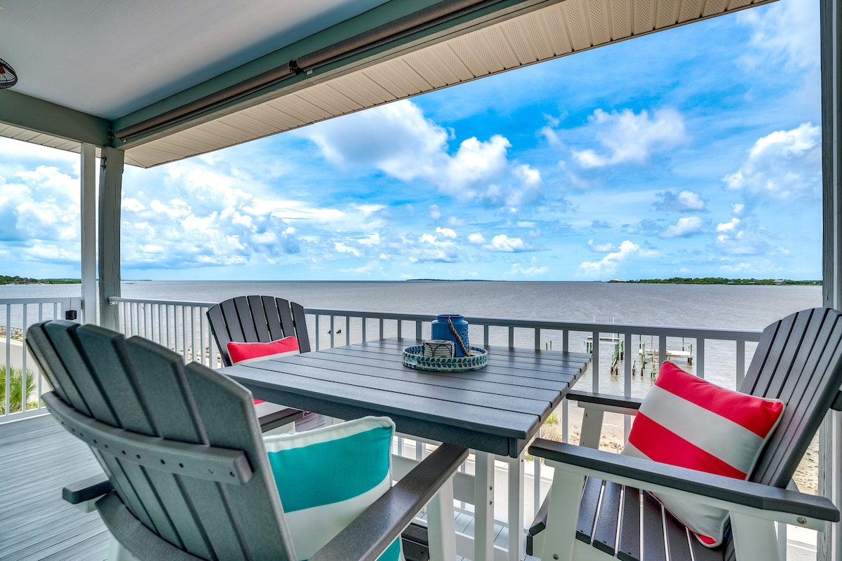A balcony is seen overlooking the expansive Gulf waters, featuring a dining table surrounded by two adirondack-style chairs. Brightly colored cushions in red and turquoise add a pop of color to the seating area, with a coastal-themed centerpiece on the table.