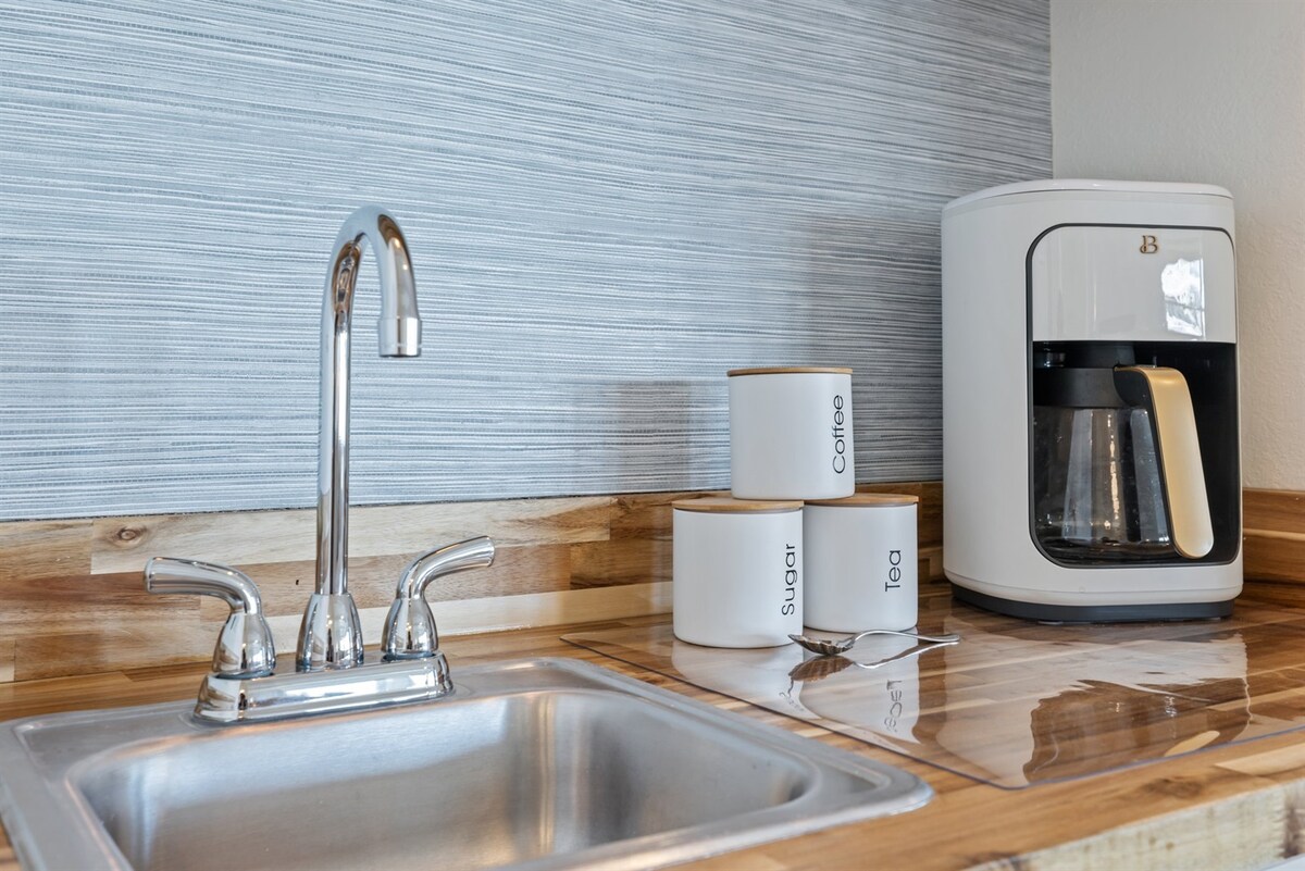 A clean and functional kitchen corner features a stainless steel sink with a polished chrome faucet. To the right, a modern coffee maker sits next to labeled canisters for sugar, coffee, and tea, all atop a smooth wooden countertop.