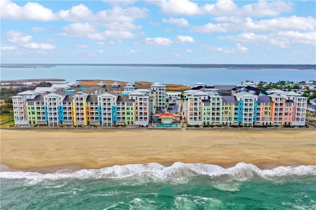 An aerial view captures the colorful beachfront condominium complex, showcasing a mix of pastel shades. The building's proximity to the sandy beach and serene waters emphasizes its coastal charm. The expansive landscape highlights the tranquil atmosphere of the surrounding environment.