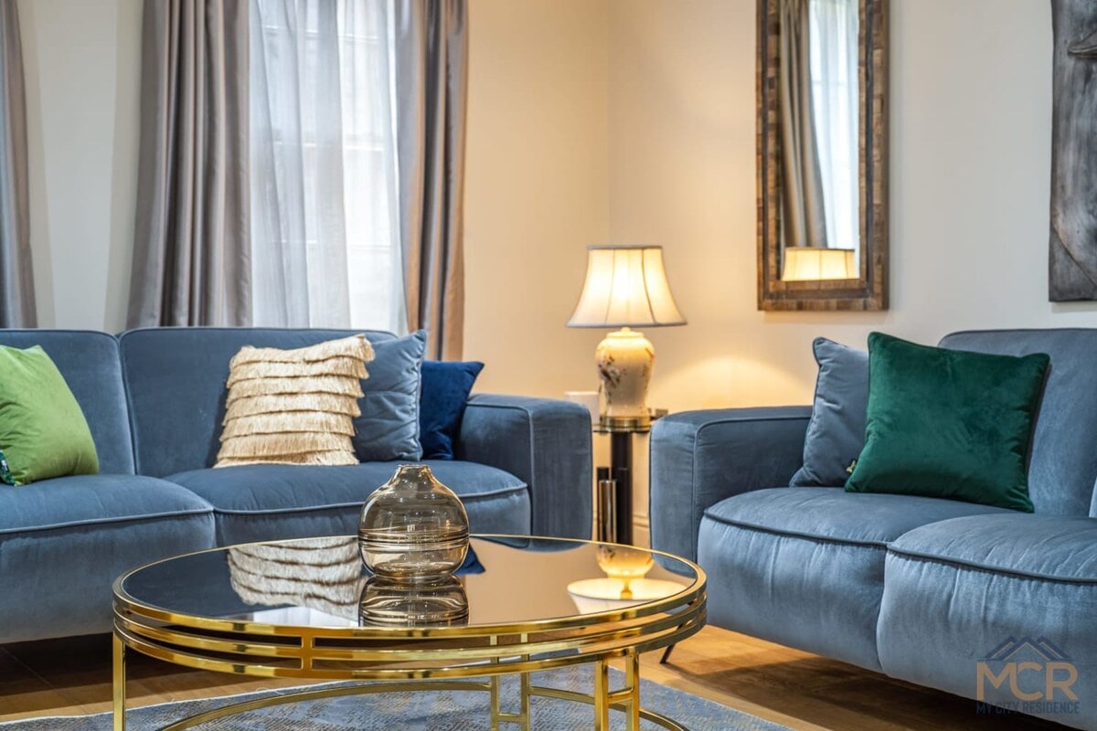 A comfortable living room features two plush sofas adorned with decorative pillows in vibrant colors. A stylish glass coffee table sits at the center, complemented by a soft rug. Natural light filters through the curtains, creating a welcoming ambiance.