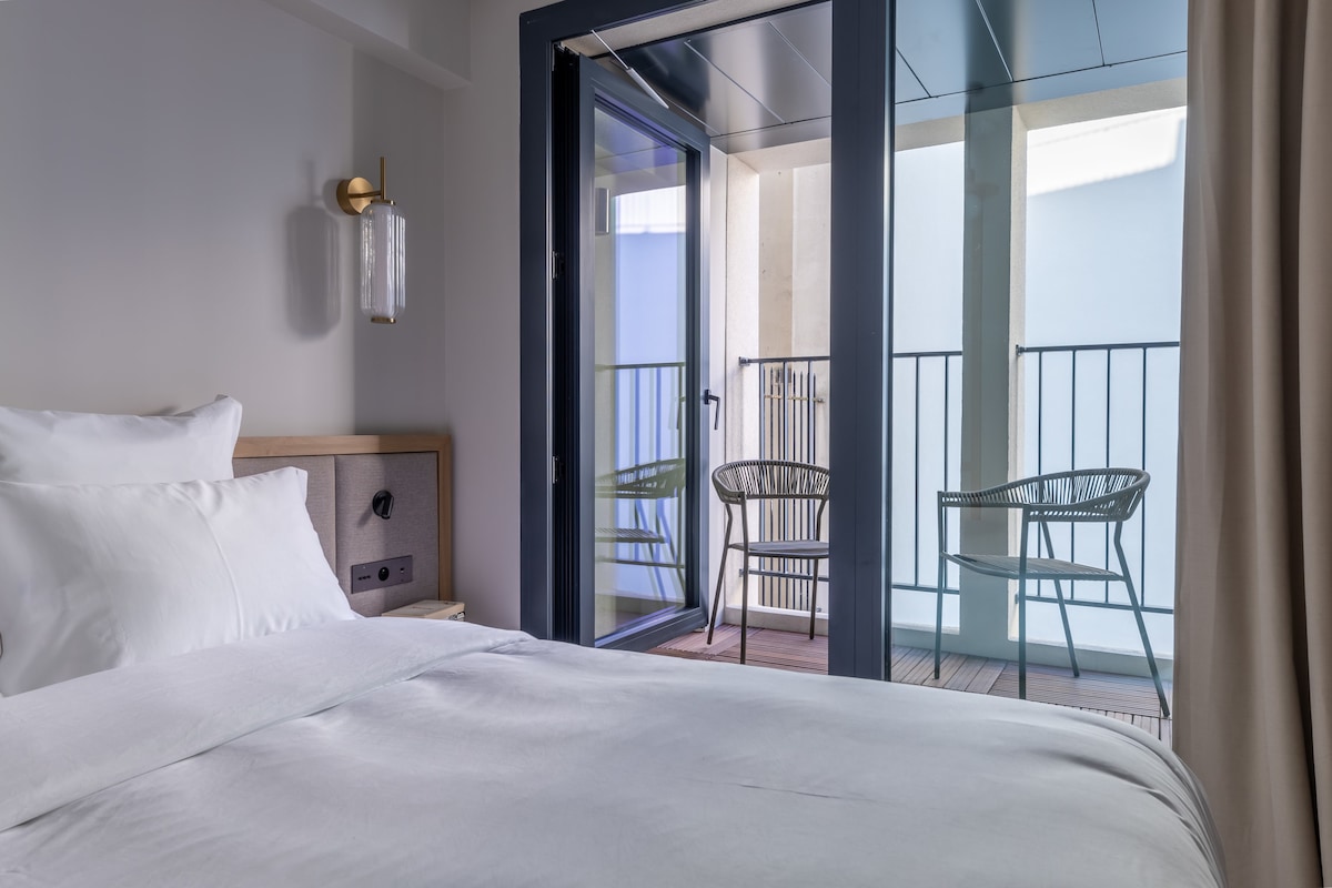 A spacious bedroom features a comfortable bed with soft white linens. Glass doors lead to a balcony with two stylish chairs, allowing natural light to fill the room. Neutral tones are complemented by a modern wall sconce, adding a touch of elegance.